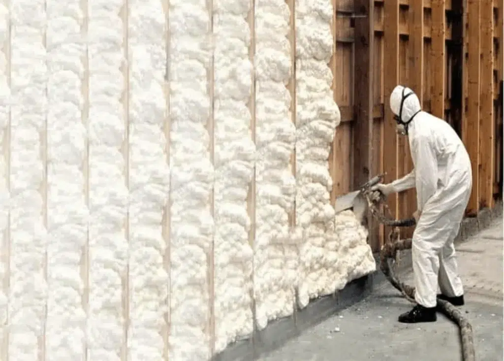 Installer in a white protective suit spraying foam insulation onto a wood-framed wall